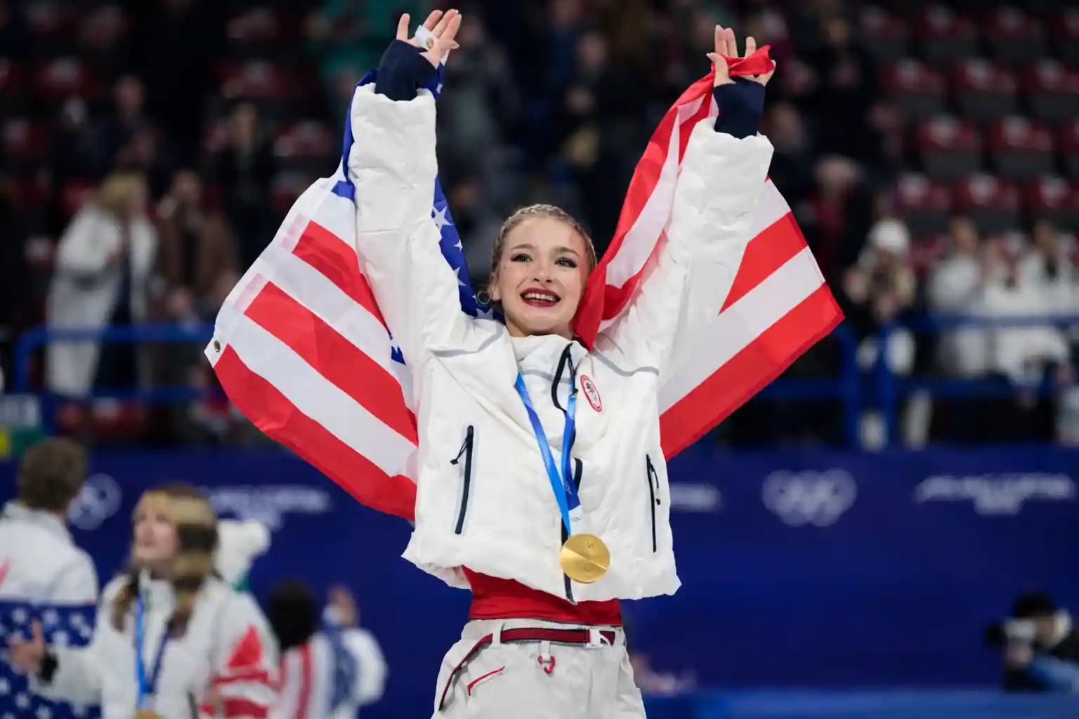 Amber Glenn celebrating with a gold medal and the American flag after winning the team event at the 2026 Winter Olympics.