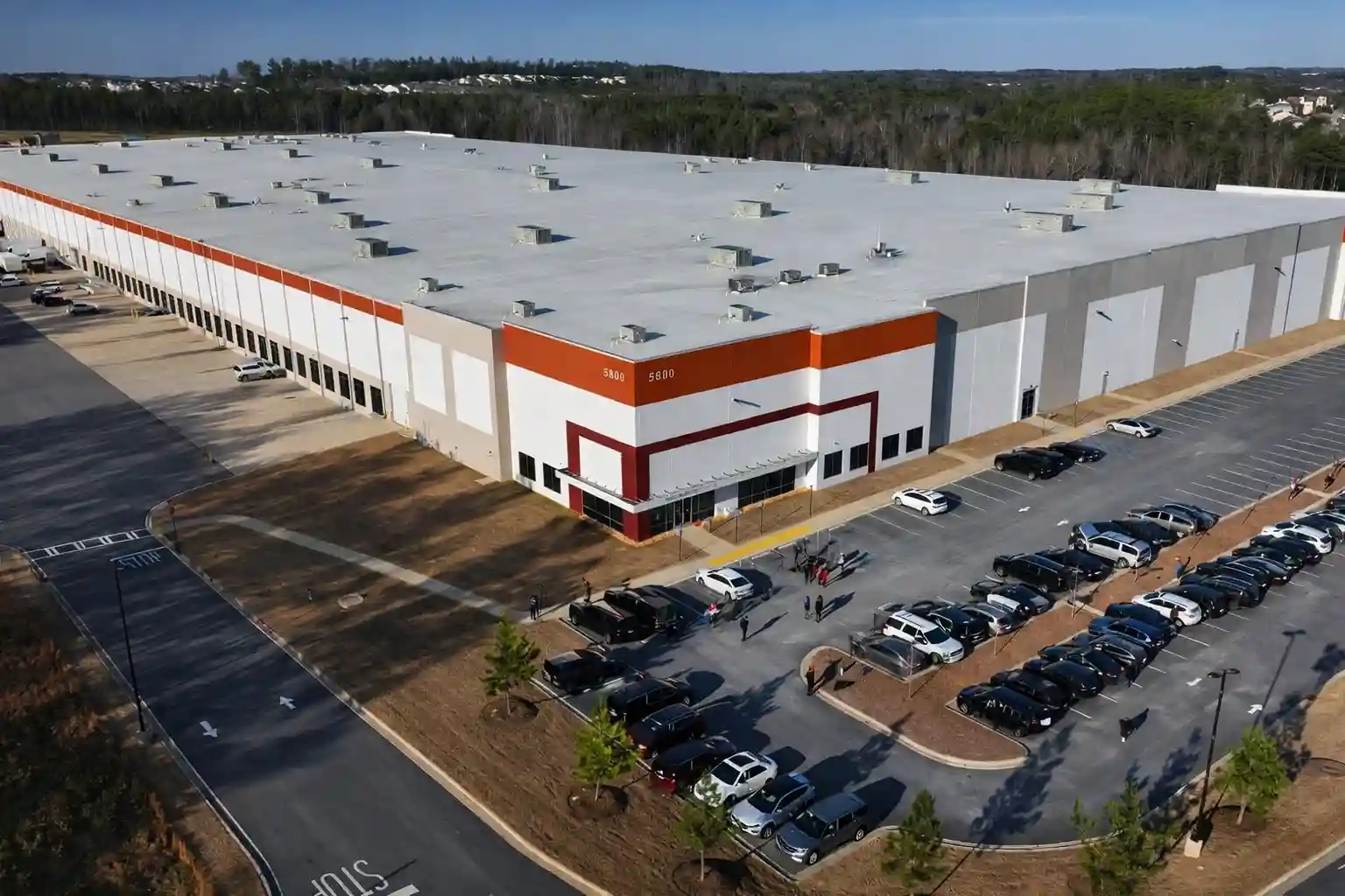 An aerial view of a large warehouse with a parking lot, showing vehicles parked near the building entrance.